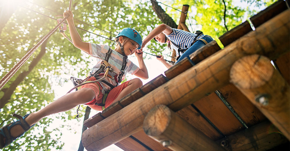Zwei Kinder in Kletterausrüstung klettern bei sonnigem Wetter auf einem Holzgerüst herum.